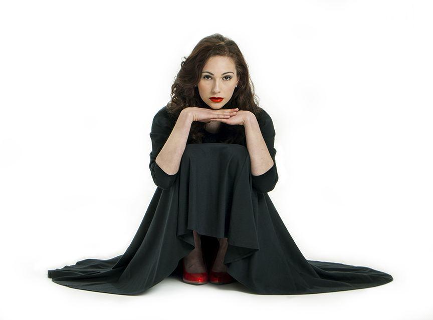 Woman in a black cape dress and red heels, chin resting on her hands against a white seamless