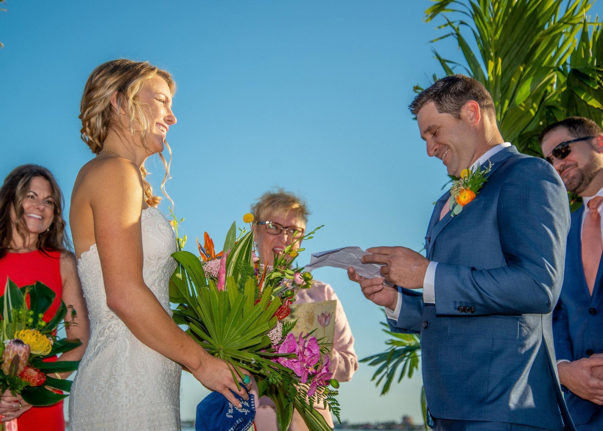 Bride in a strapless lace gown smiling as the groom reads vows beside tropical bouquets and blue sky